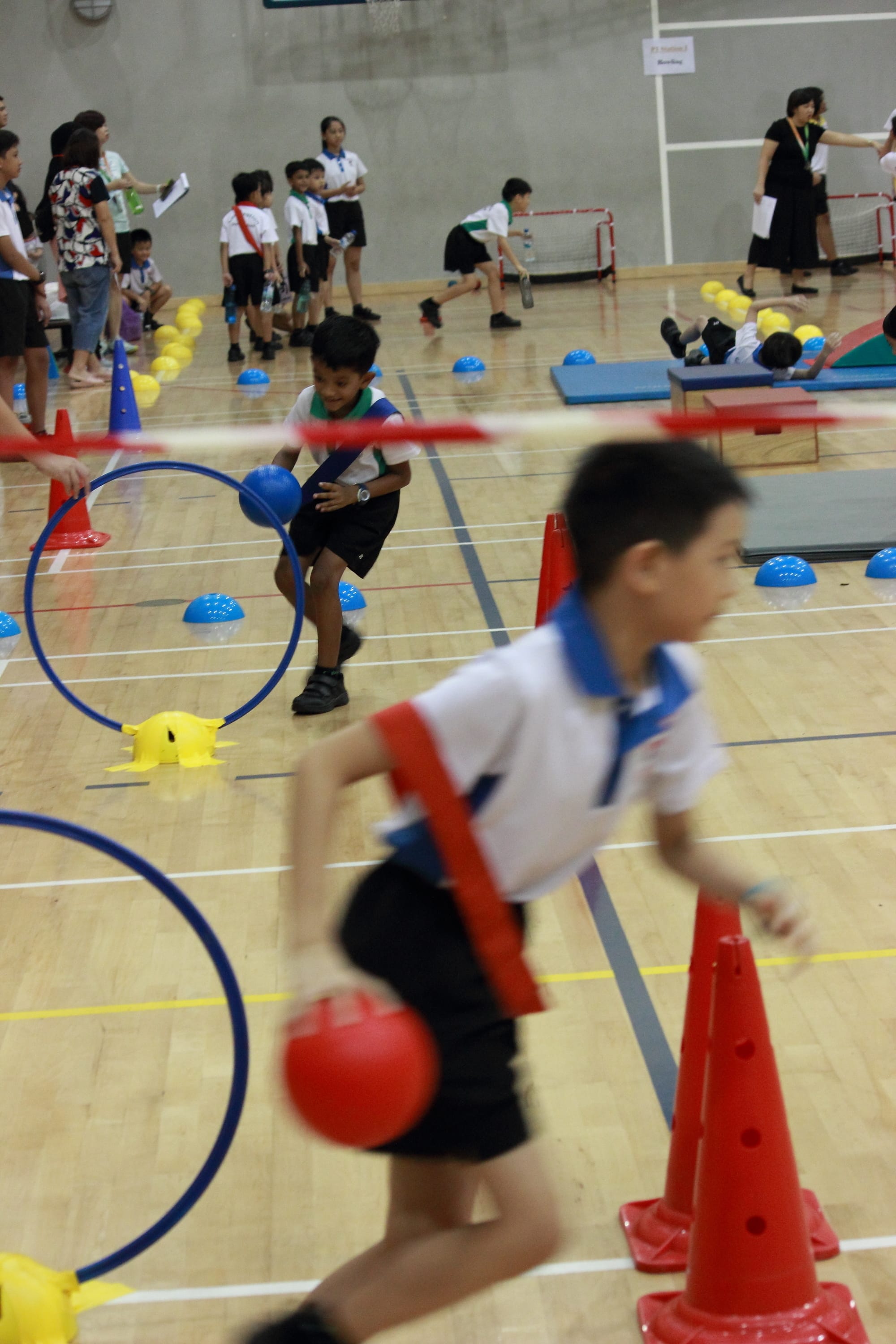 Children in uniforms do an indoor sports obstacle course with cones, hoops, balls, and mats.